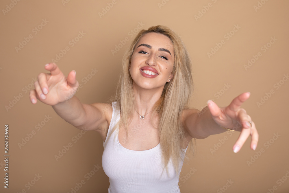 Friendly looking blonde female pulls hands towards camera, gives warm hug, smiles joyfully, going to cuddle with best friend, wears white casual outfit, isolated over beige background. Friendly looking blonde female pulls hands towards camera, gives warm hug, smiles joyfully, going to cuddle with best friend, wears white casual outfit, isolated over beige background.