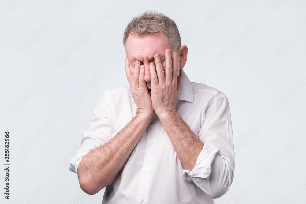 Senior man in white shirt covering his face with both hands Senior man in white shirt covering his face with both hands