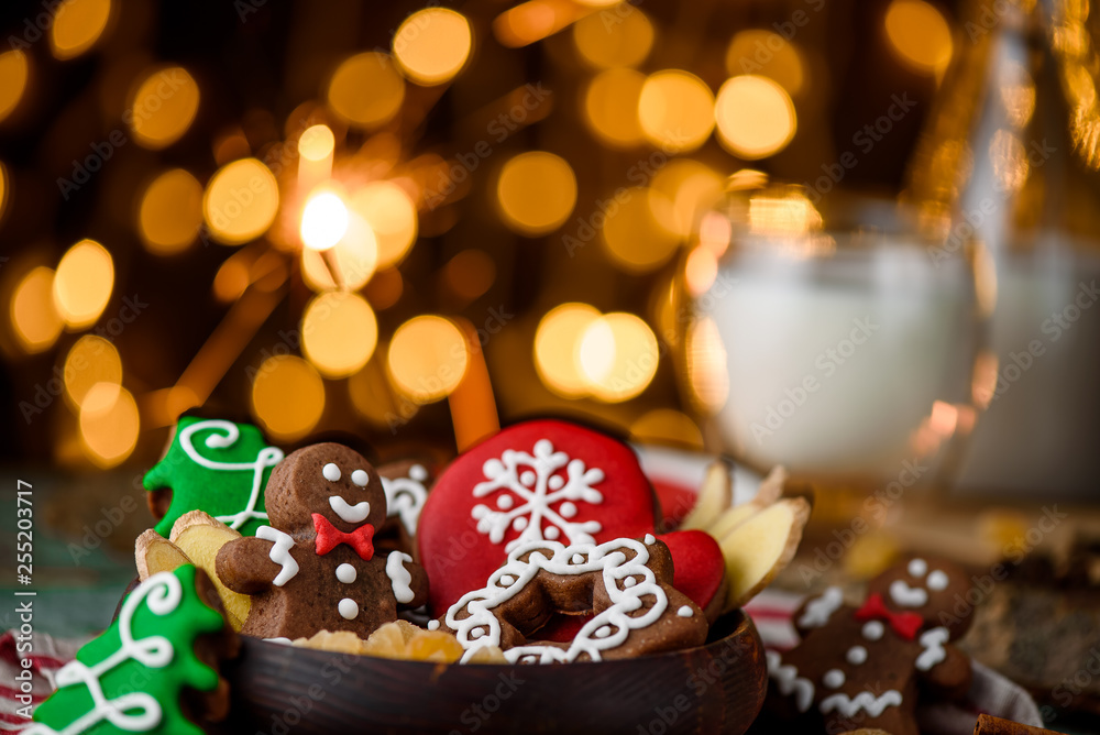 Gingerbread mittens, little man, snowflakes, christmas trees in a brown wooden plate and a bottle of milk on a green wooden on the background of blurred garland. close up. space