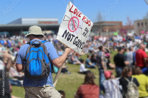 Back view of man in shorts and teeshirt with hat and backpack at gun control rally with a sign that says LOVE KIDS - Ban ARs not guns in front of blurred out crowd and stadium © Susan Vineyard 