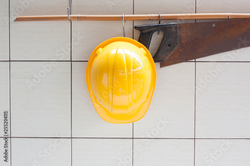 Yellow hard hat and hand saw hanging on tile wall.