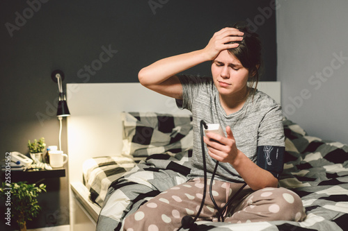 The topic of high blood pressure is hypertension disease. 22 year old young Caucasian woman uses an automatic tonometer to check pressure at home in the bedroom on the bed in home clothes in evening photo