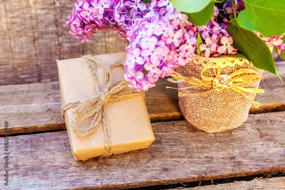 Floral decor elements. Small gift box with presents wrapped craft brown paper with bouquet of flowers beautiful smell violet purple lilac in vase on rustic wooden background. Copy space Floral decor elements. Small gift box with presents wrapped craft brown paper with bouquet of flowers beautiful smell violet purple lilac in vase on rustic wooden background. Copy space