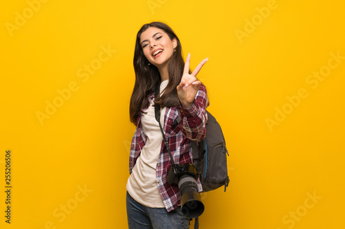 Photographer teenager girl over yellow wall smiling and showing victory sign