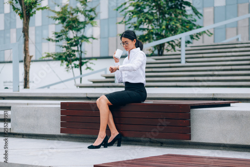 Asian businesswoman in eyeglasses looking at her wristwatch for checking the time while drinking coffee sitting in front office building background- woman office worker and businesswoman concept
