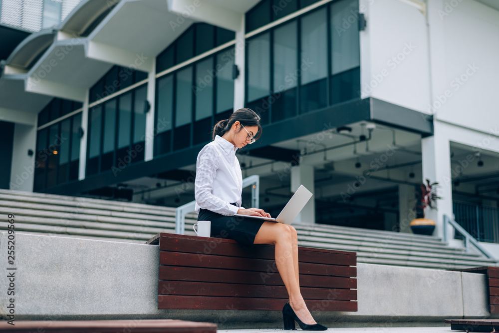 Asian businesswoman in eyeglasses using laptop working about business and finance with a cup of coffee sitting in front office building background- woman office worker and businesswoman concept