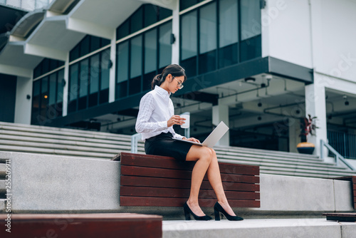 Asian businesswoman in eyeglasses using laptop working about business and financial while drinking coffee sitting in front office background- woman office worker and businesswoman concept