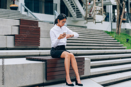 Asian businesswoman in eyeglasses using smartphone read and text message while looking at her wristwatch for checking time sitting in front office building background- woman office worker concept