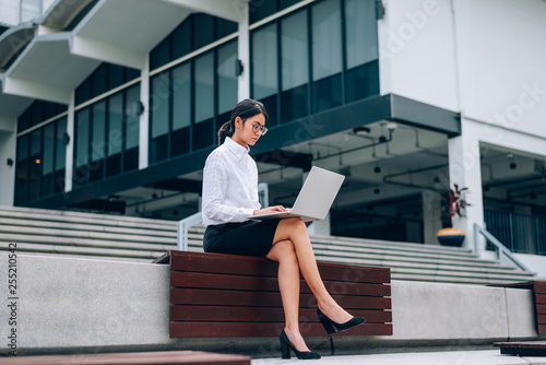 Busy Asian businesswoman in eyeglasses using laptop working mode about business and financial sitting in front office building background- woman office worker and businesswoman concept