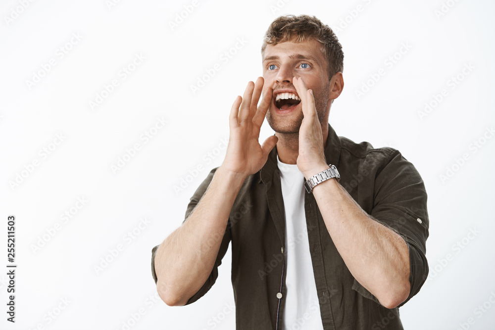 Happy charismatic young blond man with bristle and blue eyes shouting out loud holding hands near opened mouth as calling into distance smiling looking with excitement and joy, posing over gray wall Happy charismatic young blond man with bristle and blue eyes shouting out loud holding hands near opened mouth as calling into distance smiling looking with excitement and joy, posing over gray wall