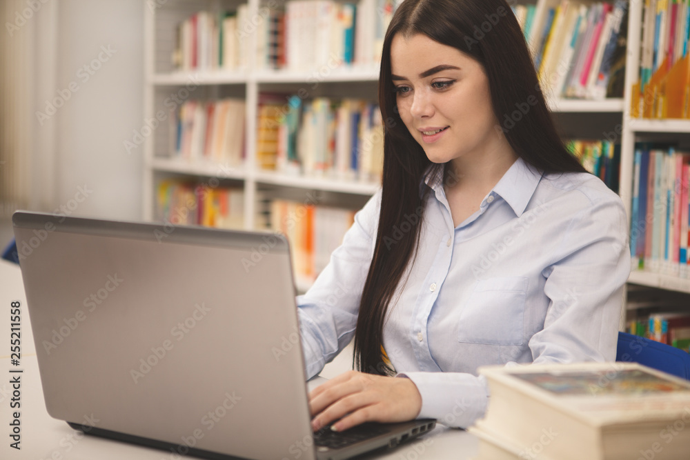 Attractive confident female student smiling, typing on her laptop, working at college library. Happy beautiful woman enjoying e-learning program. Intelligence, international education concept Attractive confident female student smiling, typing on her laptop, working at college library. Happy beautiful woman enjoying e-learning program. Intelligence, international education concept