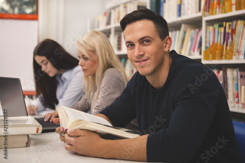 Handsome young man smiling to the camera, while studying with his classmates at the college library. Cheerful male student reading a book, preparing for exams at university. Teamwork, youth concept