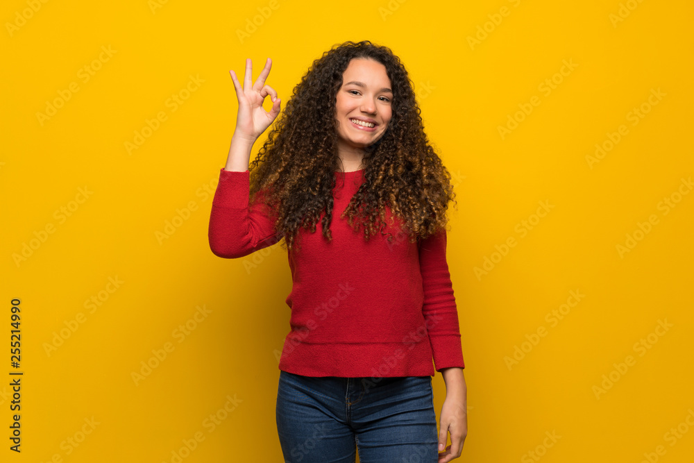 Teenager girl with red sweater over yellow wall showing ok sign with fingers Teenager girl with red sweater over yellow wall showing ok sign with fingers