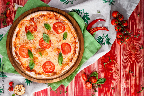 tasty traditional margarita pizza on a round wooden board on a red wooden background, decorated with napkins, chili pepper and cherry tomatoes. close-up. top view. for menu