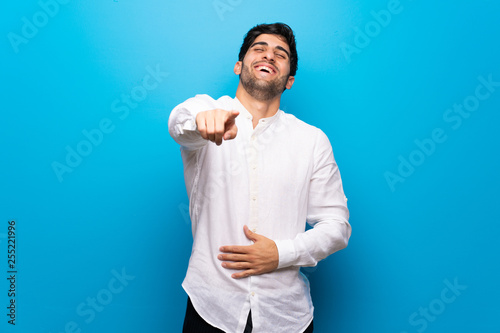 Young man over isolated blue wall pointing with finger at someone and laughing a lot