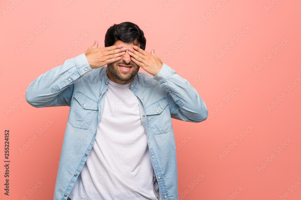 Young man over pink wall covering eyes by hands Young man over pink wall covering eyes by hands