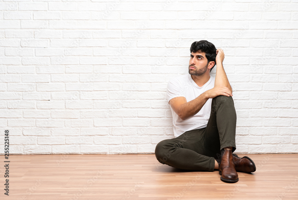 Young man sitting on the floor having doubts while scratching head Young man sitting on the floor having doubts while scratching head