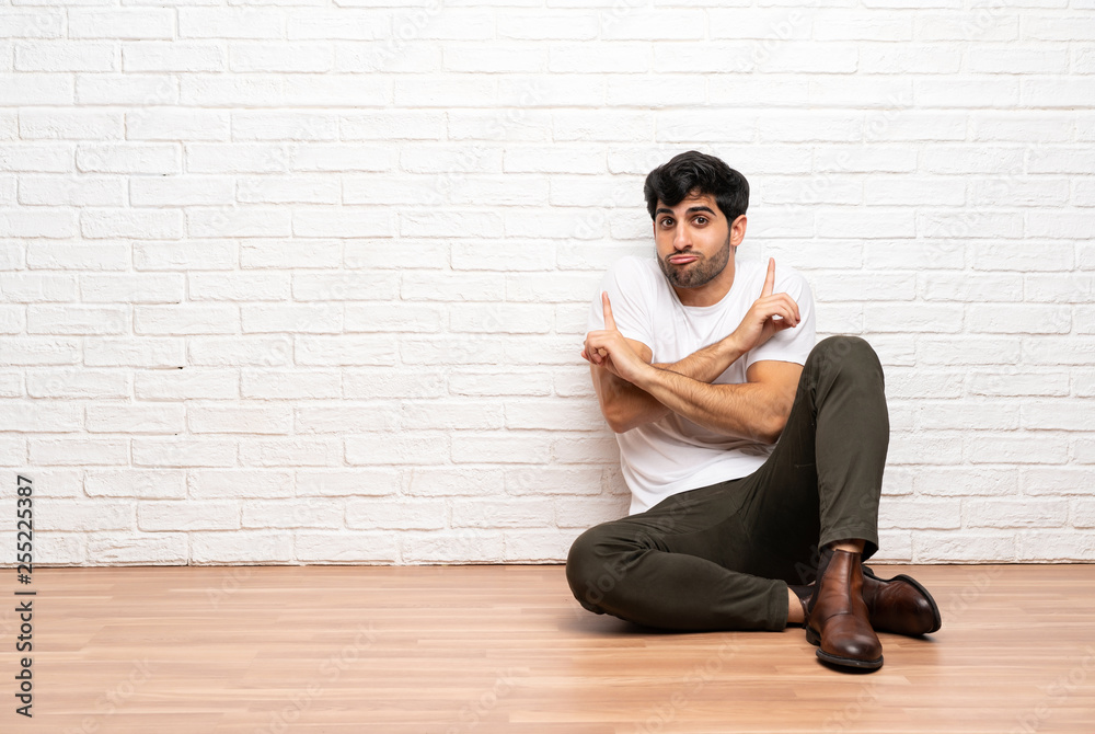 Young man sitting on the floor pointing to the laterals having doubts Young man sitting on the floor pointing to the laterals having doubts
