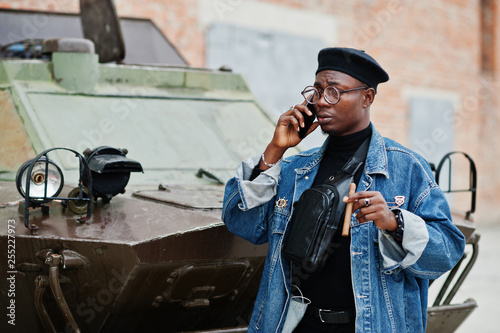 African american man in jeans jacket, beret and eyeglasses, with cigar posed against btr military armored vehicle, and speaking on mobile phone.