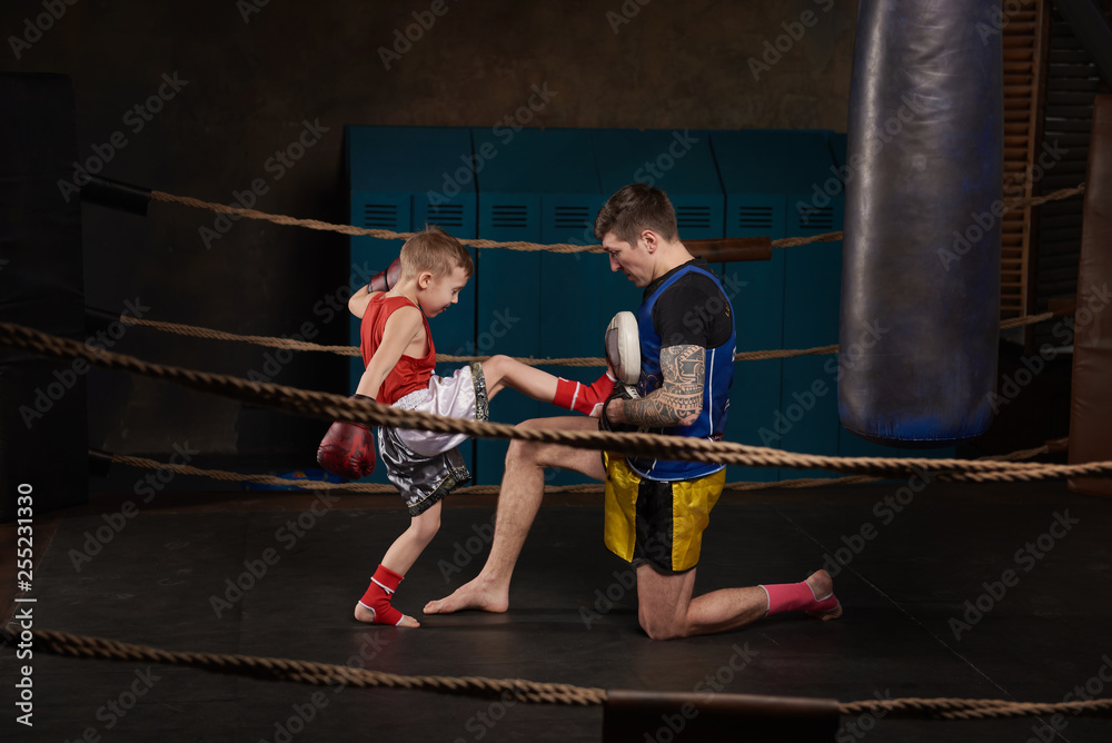 Trainer teaching kid how to hit punches. Kid wearing boxing gloves and head guard training with his coach inside a boxing ring