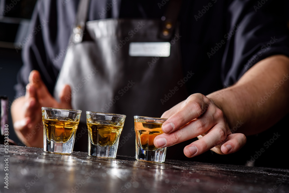 Bartender in a dark leather apron puts three glasses of whiskey on a dark wooden bar in a nightclub. Close-up. Spa ce