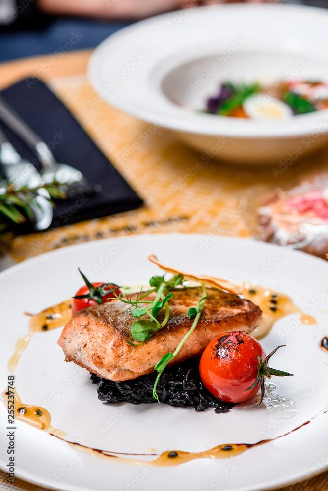 Beautiful serving of roasted salmon fillet and risotto with cuttlefish ink on a white plate with a signature sauce and Cherry tomatoes in a restaurant. close up