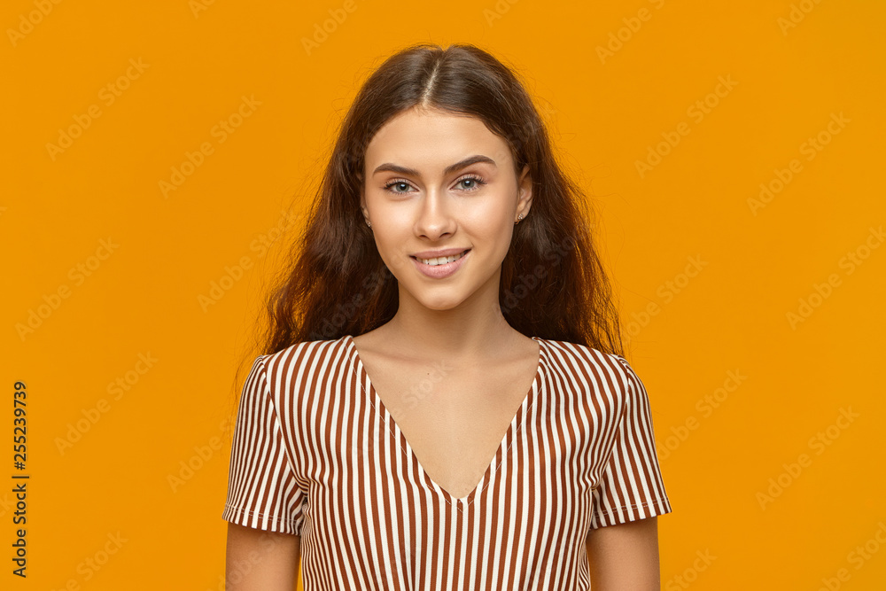 Horizontal image of gorgeous beautiful young Caucasian female in romantic summer dress posing at orange studio wall with broad cheerful smile, being in good mood, having carefree facial expression Horizontal image of gorgeous beautiful young Caucasian female in romantic summer dress posing at orange studio wall with broad cheerful smile, being in good mood, having carefree facial expression