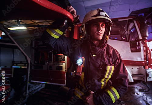 Portrait of a handsome fireman wearing a protective uniform with flashlight included standing in a fire station garage and looking sideways