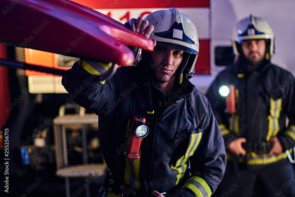 Fototapeta premium Two firemen wearing a protective uniform standing next to a fire truck. Arrival on call at night time