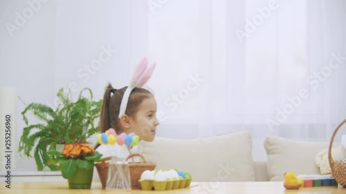 Joyful playing girl with bunny ears on his head is hiding under the wooden table by a slow pace. Girl is dowing down slowly hiding herself. photo