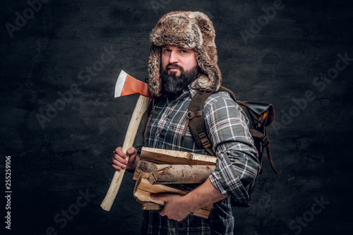 Portrait of a bearded woodcutter with a backpack dressed in a plaid shirt and trapper hat holding firewood and ax. Studio photo against a dark textured wall photo