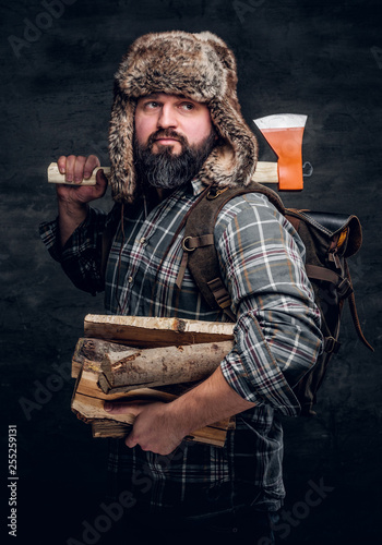 Portrait of a bearded woodcutter with a backpack dressed in a plaid shirt and trapper hat holding firewood and ax. Studio photo against a dark textured wall