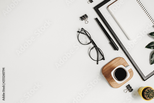 Flat lay office workspace with blank clip board, office supplies, pen, cactus, green leaf, coffee cup on a wooden stand and eyeglasses on white background. Top view minimal mock up template concept