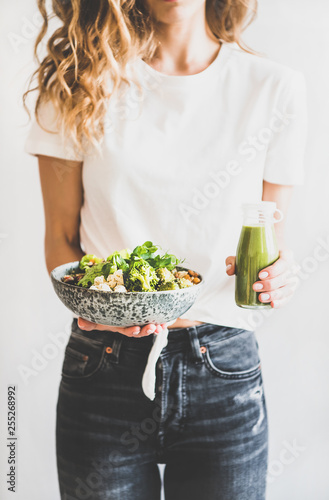 Healthy dinner, lunch. Woman in jeans standing and holding vegan superbowl or Buddha bowl with hummus, vegetable, fresh salad, beans, couscous and avocado and green smoothie in bottle photo