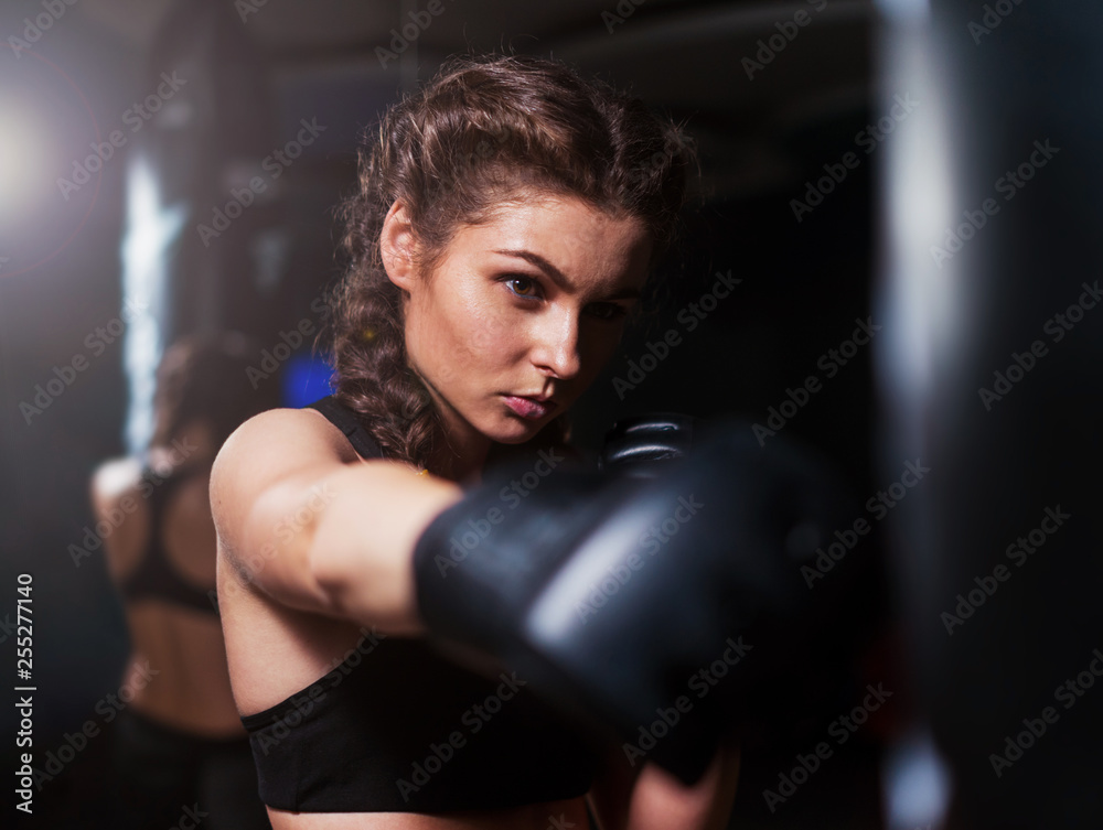 Young fighter boxer fit girl wearing boxing gloves in training with heavy punching bag in gym. Low key image. Woman power. Moment of punch