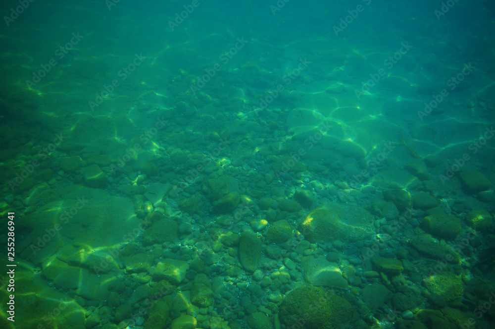 Gravel bottom in shallow green sea with areas of larger rocks and boulders and reflections from surface.