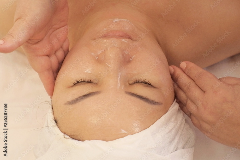 Close-up. Portrait of a woman of Asian appearance receiving a Wellness massage of the face. A woman with closed eyes lies on the couch at the beautician. Shooting from above