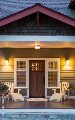 Front porch and entry of traditional Craftsman style home at  twilight dusk with windows, wooden front door, exterior lighting and Adirondack chairs