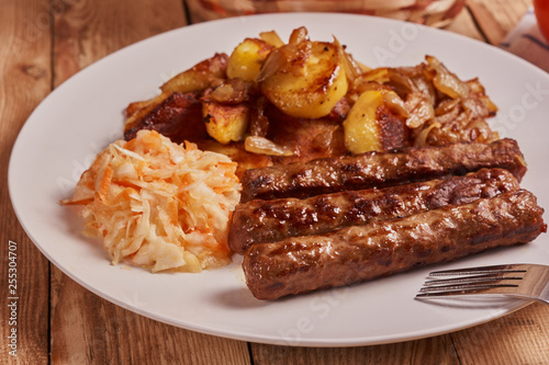 Serbian cevapi, cevapcici, Balkan minced meat kebab on a white plate with marinated cabbage, fried potatoes and fried onions next to fork on a wooden background