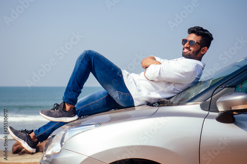 success traveler driver indian model male in a white shirt freelancer smiling and posing seacost .Handsome bearded man is standing near car freelancing on the beach © yurakrasil
