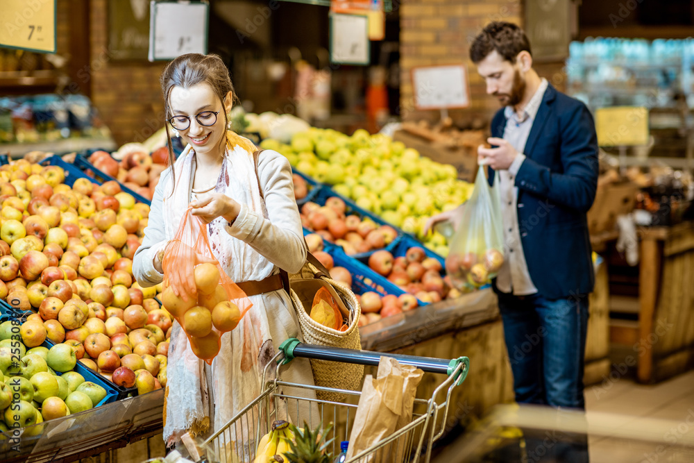 Woman packing apples into the eco bag with man holding plastic bag on the background in the supermarket Woman packing apples into the eco bag with man holding plastic bag on the background in the supermarket