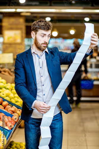 Portrait of an elegant man with shocked emotions holding very long shopping list while buying food in the supermarket photo
