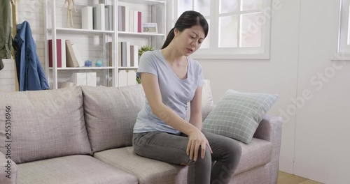 slow movement woman having knee pain sitting in sofa at home. young people osteoarthritis joint after sport breaks and sprains. asian lady moving painful legs massage.