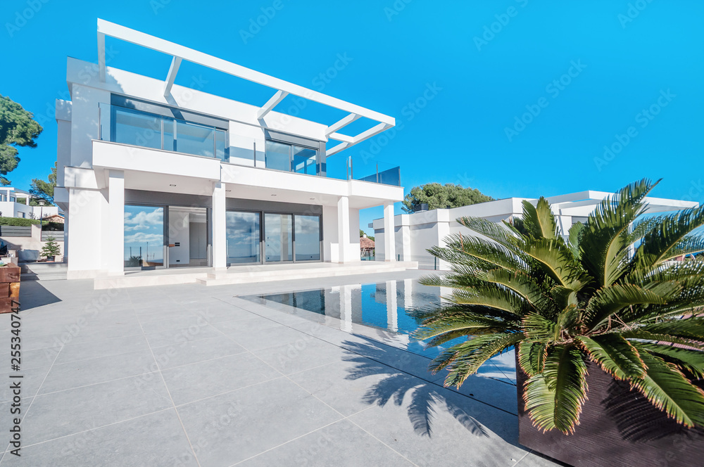 Luxury modern white house with large windows overlooking a Mediterian landscaped garden with palm trees and blue swimming pool. High tech style villa. Vacation home or hotel. Modern loft design.ees a