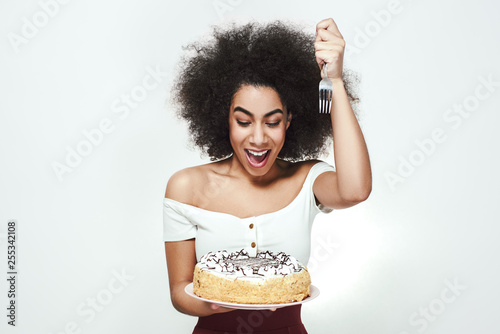 I will eat it right now! Excited cute afro american woman with curly hair is holding a fork in one hand and birthday cake in another, going to eat some piece of cake. photo