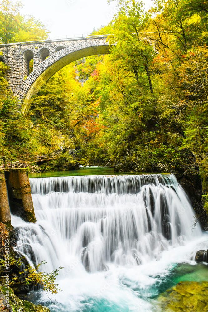 Autumn forest colors with turquoise waterfall and old rocky train bridge in natural park of Vintgar river gorge Slovenia