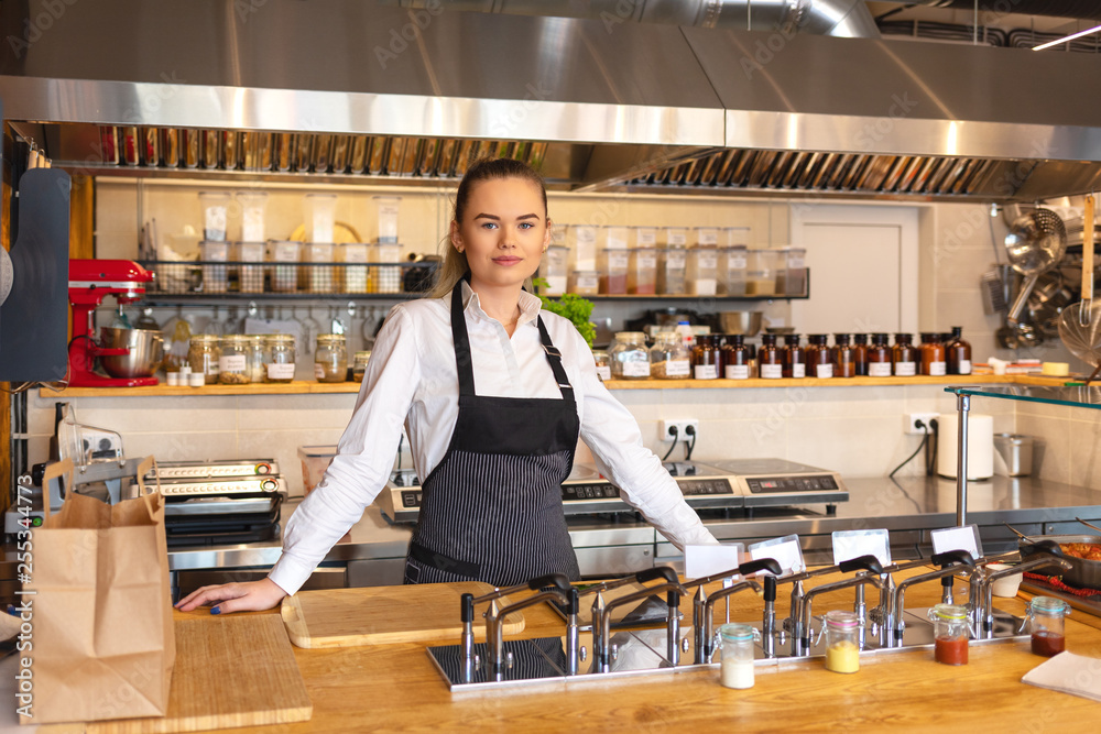 Portrait of young woman standing behind kitchen counter in small eatery – restaurant owner wearing apron looking confident at camera smiling