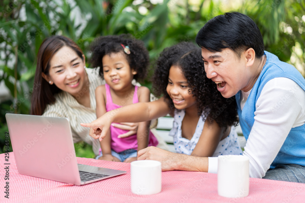 Happy family checking their social media or video call on a laptop computer as they relax together on backyard. parents or Volunteers take care of orphans pretty little daughter in child care center Happy family checking their social media or video call on a laptop computer as they relax together on backyard. parents or Volunteers take care of orphans pretty little daughter in child care center