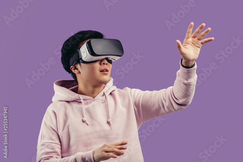 Amazed positive chinese male gamer wearing virtual reality goggles headset, vr box, posing isolated in studio over violet background. Connection, technology, new generation, progress concept.