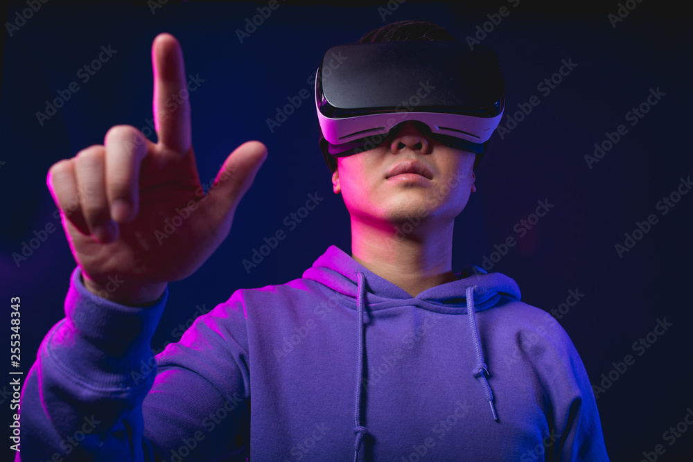 Joyful Japanese youngster dressed in sport hoodie, wearing virtual reality helmet on dark blue background is pointing to camera direction. Smartphone using with virtual reality goggles. Joyful Japanese youngster dressed in sport hoodie, wearing virtual reality helmet on dark blue background is pointing to camera direction. Smartphone using with virtual reality goggles.